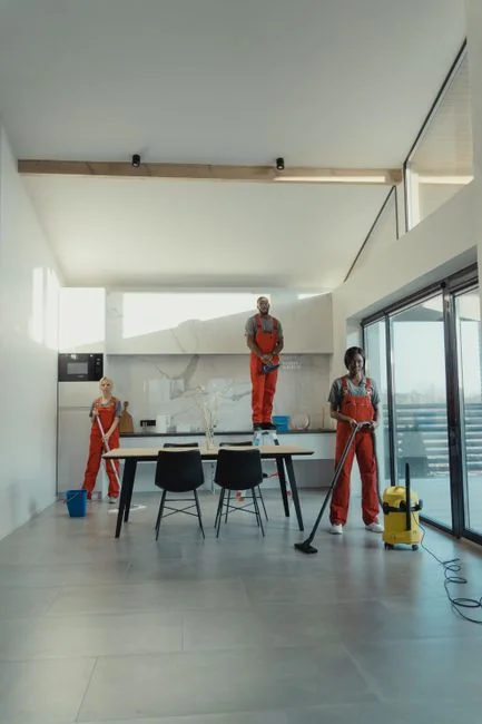 Two women in work clothes cleaning a modern living room using a vacuum and mop.